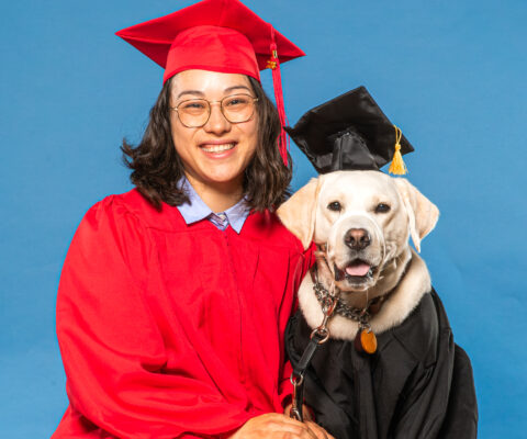 women in graduation gown sits next to dog in graduation gown